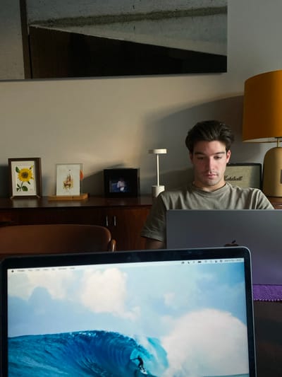 This image depicts an indoor scene, likely a home or office, situated in Lordelo do Ouro e Massarelos, Portugal. In the foreground, the top half of a laptop screen is visible, displaying a wallpaper of a surfer riding a large blue wave under a partly cloudy sky, with the macOS menu bar showing "Mon 6 Oct 18:05". Behind this, a young adult male with dark hair, wearing a light-colored t-shirt, is seated and intensely focused on a silver laptop positioned directly in front of him. A purple fabric mat is visible beneath his laptop. In the background, a dark wooden cabinet/sideboard holds several items: a framed picture of a sunflower, another framed artwork, a small black digital photo frame, and a white minimalist table lamp. To the right, an orange-shaded table lamp with a brass-colored base is present, and a "Marshall" branded amplifier or speaker is partially visible behind the male's head. The wall behind the cabinet is light grey, with a large abstract art piece featuring geometric shapes in dark grey, black, and light grey mounted higher on the wall. Part of a brown chair is visible on the left side of the frame.