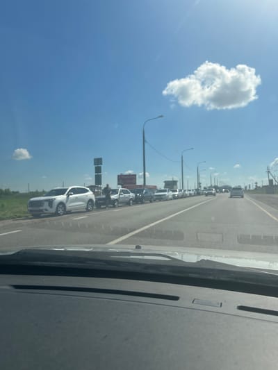 The image shows a roadside scene on a sunny day, with a clear blue sky and a few scattered white clouds. The perspective is from inside a vehicle, looking out towards the right. A long line of cars is visible on a highway, stretching into the distance. The cars are mostly white, and many appear to be SUVs or sedans. Streetlights line the side of the road, and there are some buildings and signs in the background. One sign, partially obscured, appears to have Russian text. The overall impression is of a traffic jam or a busy road during daylight hours. The sun is bright, suggesting it's likely daytime, possibly late morning or afternoon. The road appears to be paved and well-maintained. The presence of multiple cars and the highway infrastructure suggest a developed area, possibly suburban or on the outskirts of a city.