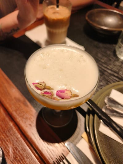 A close-up, top-down shot shows a cocktail topped with foamy white liquid, garnished with three small pink rosebuds. The drink is served in a coupe glass and rests on a dark, possibly wooden table. To the left, a portion of a person's arm with a tattoo is visible, suggesting a social gathering. In the background, a tall glass of iced coffee is on the table, along with a dark ceramic bowl. The scene appears to be in an indoor setting, possibly a restaurant or bar, given the table setting which includes cutlery and plates. The lighting is dim, with a slight warm tone. This image does not provide any specific visual cues to identify its location in Bnei Dror, Israel.
