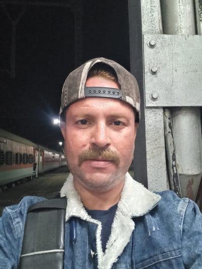 A selfie shot of a light-skinned man with a mustache wearing a denim jacket with a white sherpa collar and a baseball cap. The background is dark but suggests an elevated structure possibly a bridge or overpass. The photo appears to be taken at night in Igatpuri, India, as suggested by the context.