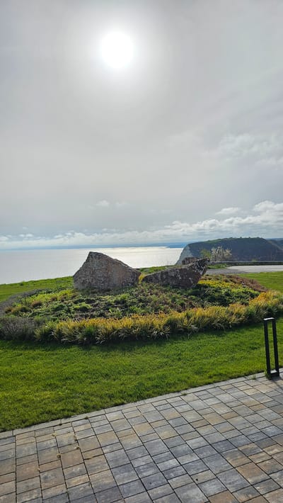 The image shows a view from a coastal area in Kavarna, Bulgaria. In the foreground, there is a paved area, bordered by a strip of green grass interspersed with yellow plants. Adjacent to this grass is a cliff face overlooking a body of water, likely the Black Sea. Beyond the sea, the outline of another landmass or cliff is visible. In the sky, the sun is shining brightly, with some cloud cover present. The image is taken during daytime and the overall scene is of a natural landscape with man-made elements along the coast.