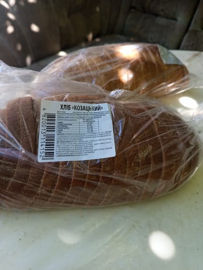 This media file features a close-up view of two loaves of dark bread, likely rye or a mixed grain, individually packaged in clear plastic. The bread appears to be sliced, with visible crusts and a dark, dense crumb. The packaging is slightly wrinkled, suggesting the bread is fresh.
A prominent label is affixed to the front loaf, providing product information in Ukrainian. The label indicates the product is "ХЛІБ «КОЗАЦЬКИЙ»" (Bread "Cossack"). It details nutritional information per 100g, including energy value (226 kcal), fats (1.1g), carbohydrates (47g), and proteins (7.1g). Storage instructions recommend temperatures between +6°C and +28°C. The manufacturer, TDV "Eychdi Bakeri Ist," and its address in Kyiv, Ukraine, are listed, along with production facility addresses in Zaporizhzhia. A barcode and the lot number, which is linked to the "use by" date, are also present.
The background suggests an outdoor setting, possibly a table or surface outdoors, with diffused sunlight casting shadows and highlights on the packaging and the bread. The overall impression is a product photograph, likely taken in natural light. The context of Kramatorsk, Ukraine, places this image within a specific geographic and cultural setting.