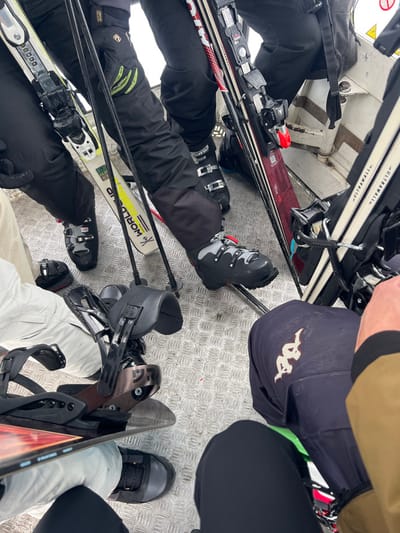 The image displays the interior of a ski lift cabin in the ski resort area of Valtournenche, Italy. The floor is constructed from diamond plate metal, and white painted metal structural elements are visible along the right side. At least four individuals are depicted, primarily from the waist down, equipped with winter sports gear. Clothing observed includes black ski pants, one pair of light-colored pants, and a dark blue pair of pants featuring a white Kappa logo on the right thigh. Multiple pairs of black ski boots are worn by individuals, alongside at least one pair of black snowboard boots. Ski equipment, including several pairs of skis with bindings (one white ski is branded "WORLD CUP," another is red, and others are black/white), and black ski poles, are positioned upright between the occupants. A snowboard with black bindings is also visible in the lower-left quadrant of the frame. The individuals appear to be seated or standing closely together, being transported with their equipment within the confined space of the cabin.