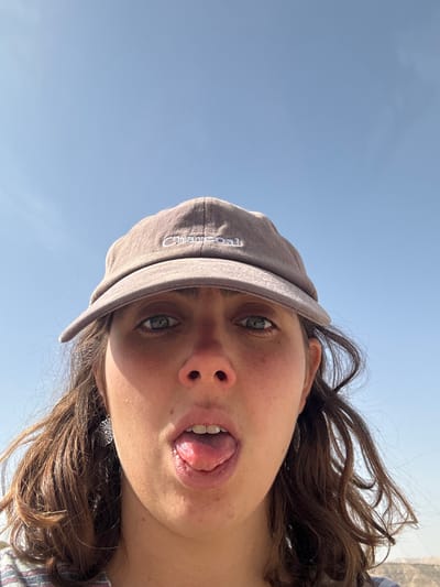 A close-up, head-on selfie, taken in the Megilot Yam HaMelakh Regional Council, Palestinian Territory, features a single female individual. She has fair skin, blue-green eyes, and brown, wavy hair. Her mouth is open with her tongue extended outwards. She wears a grey baseball cap with "Charcoal" embroidered in white on the front, and a silver-colored, intricate circular earring is visible on her left ear. The background is a clear, light blue sky, with a hazy, light-toned horizon or distant landscape faintly visible at the bottom right.