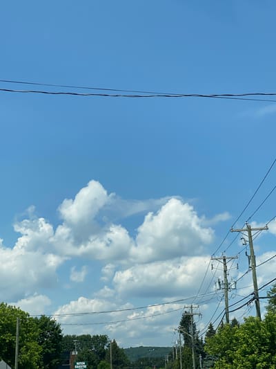 The image captures a bright, partly cloudy sky above a residential street in Saint-André-Avellin, Canada. Power lines crisscross the frame, supported by wooden utility poles. Lush green trees line the street, with one pole in the foreground bearing a green sign that reads "NATION ÉQUESTRE." The scene is devoid of people.
