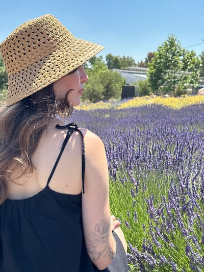 An over-the-shoulder shot captures a woman wearing a straw hat and a black dress, positioned in a field of vibrant purple lavender under a clear blue sky. The lavender field extends into the background, interspersed with patches of yellow flowers and green trees. In the distance, buildings and a greenhouse are visible, suggesting an agricultural or park setting. The woman's hair is brown and wavy, and she has a tattoo on her left arm. The image conveys a sense of warmth and natural beauty, likely taken on a sunny day in a rural or semi-rural area.
