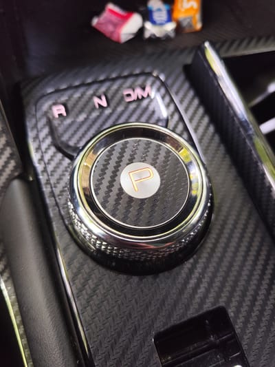 The image shows a close-up of a car's gear shifter, which is set to "Park" (indicated by the "P"). The gear shifter is surrounded by a carbon fiber trim. In the background, there are some small, colorful objects, possibly candy wrappers or small toys. The gear selection indicator lights up "R", "N", and "D" in pink. The setting appears to be indoors, likely the interior of a vehicle. There is no indication of Petropavl City Administration, Kazakhstan.