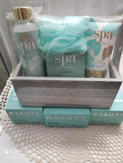 This is a photograph of a spa gift set containing a bottle of bath and shower gel, a bar of hand soap, and a tube of body lotion. The products are branded "Spa Elements" and have a pale green and gold color scheme with botanical-inspired designs. A light blue mesh bath pouf is placed on top of the products. The gift set is housed in a rustic wooden box with a weathered grey finish. Below the wooden box are three teal-colored boxes, two of which are partially visible, also branded "BEAUTY". One of these boxes appears to be for a "SILK FACIAL MASK" and another for "FACE CREAM NATURAL SERIES".

The items are arranged on a white crocheted doily. The background is blurred but suggests an indoor setting, possibly a bathroom or dressing area. The lighting appears to be artificial, casting soft shadows.

Visible text includes product branding such as "Spa Elements", "BATH & SHOWER", "HAND SOAP", "BODY LOTION", "BLOSSOM & TEA", and "BEAUTY". There is also a timestamp at the bottom right of the image: "25.06.2026 09:30". In the bottom left corner, text indicates the device used to take the photo: "XIAOMI 11 LITE 5G NE". The overall impression is a clean, spa-like aesthetic, suggesting relaxation and self-care. There are no people visible in the image. The setting appears to be a domestic indoor environment.