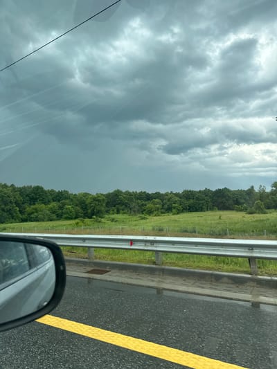The image depicts a view from a vehicle traveling along a wet, grey asphalt road marked with a bright yellow line. The road is bordered by a metal guardrail and a grassy verge. Beyond the verge, a dense line of lush green trees forms a thick forest. The sky above is overcast with heavy, dark grey clouds, suggesting impending or ongoing rain. The overall scene conveys a sense of a somber, possibly stormy, day. The image appears to be taken while the vehicle is in motion, as indicated by the slight blur and the reflection in the side-view mirror. No people are visible in the frame, and no text is discernable. The setting is rural, with the forest and open grassy areas suggesting a location outside of a city. The weather conditions are clearly the most prominent feature, with the dramatic cloud cover dominating the upper portion of the image and the wet road confirming recent or current precipitation.