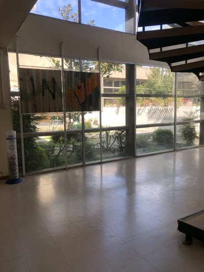 The image shows the interior of a building with a large window overlooking a courtyard. There are two abstract paintings hanging on the wall. The courtyard is filled with green shrubs and trees, and the window is reflecting the bright sky outside. There are several white ceramic tiles on the floor, some of which have scuff marks. There is a blue recycling bin in the corner of the room. The photo was taken in Agia Paraskevi, Greece. It is located in the 2nd floor of a building on the corner of 21st May and Leof. Kifisou street. The photo was taken from inside the building, looking out at the courtyard. The floor is tiled and the walls are painted a light cream color. The window is made of glass panes, and the metal bars on the outside of the window create a grid pattern. A metal staircase is visible on the right side of the photo. The courtyard is visible through the window. There are several trees and shrubs growing in the courtyard. The courtyard is surrounded by a low white wall. There are no people or other objects in the photo. The photo was taken during the day, and the sun is shining. The photo is taken in Agia Paraskevi, Greece. The building is located at 37.999, 23.82 coordinates.