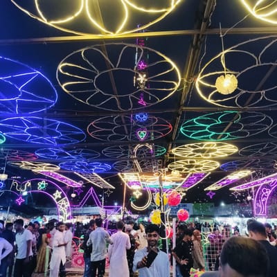 This is a vibrant nighttime photograph capturing a bustling outdoor market or festival in Charfasson, Bangladesh. The scene is illuminated by an elaborate canopy of colorful LED string lights, arranged in various circular, floral, star, and heart patterns across the overhead structure. The lights display a spectrum of colors including yellow, blue, green, red, pink, and purple, creating a festive atmosphere.

A large crowd of people, predominantly young men dressed in both casual and traditional attire like panjabis, fills the space below the lights. Many are observing their surroundings, while one man in the foreground smiles at the camera while holding a smartphone. Bubbles are visibly floating through the air, adding a whimsical touch to the gathering, and red and yellow spherical balloons are also part of the decor. In the background, market stalls or shops are discernible, indicating a commercial aspect to the event.

The overall mood appears lively and celebratory, enhanced by the bright, decorative lighting and the gathering of people. Visible text from signs includes "প্রাণিজ্য ও", "বাস মারি", "২০ইড ইং চরফ্যাশন মাস" (likely referring to Charfasson and a date/month), "সার্বিক", "টপটি", and "CLEMON", providing context to the location and possibly sponsors or vendors. The image clearly depicts an evening event, with the dark sky serving as a backdrop for the illuminated decorations.