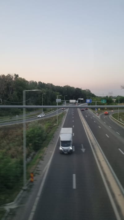 The image depicts a highway scene in the vicinity of Jilava, Romania. A white delivery van is traveling in the right lane of the highway, which is divided by a concrete barrier and white lane markings. The sky above is a pale blue, transitioning to a lighter shade near the horizon, indicating it is either early morning or late evening. On the left side of the highway, there is a wooded area with dense green trees. Several other vehicles, including cars and trucks, are visible in the distance on both lanes of the highway. Road signs are present, with one blue sign in the distance indicating directions. The overall scene is one of typical highway traffic flow under a soft, ambient light.