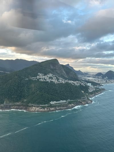 This aerial image captures a panoramic view of the coastline and urban landscape of Rio de Janeiro, Brazil, under a heavily clouded sky. The scene is dominated by a rugged, verdant mountainous terrain meeting the Atlantic Ocean.
In the foreground and midground, a dense, dark green forested mountain slope descends towards a rocky coastline. A multi-lane coastal road, featuring sections of bridges and tunnels, hugs the base of the mountain, separating it from the turbulent, teal-colored ocean. Waves with white foam crash against the shore. On the middle slopes of the foreground mountain, a dense agglomeration of small, light-colored residential structures, characteristic of a favela, is visible amidst the vegetation. A prominent white building complex with multiple interconnected sections sits on a leveled area near the coastal road.
The ocean surface exhibits subtle linear disturbances, possibly currents or wakes, extending parallel to the coastline in the lower right portion of the image.
In the background, further along the coast and inland, the sprawling urban landscape of Rio de Janeiro unfolds. Multiple mountain peaks, also densely covered in green foliage, recede into the distance. On one of these distant peaks, the iconic Christ the Redeemer statue is faintly discernible. Communication towers are visible on other more distant mountain ridges. A large body of water, likely the Lagoa Rodrigo de Freitas or Guanabara Bay, is visible behind the immediate coastal developments, surrounded by a dense array of high-rise buildings and city infrastructure.
The sky is largely overcast with thick, grey clouds, although patches of lighter blue and hints of a warm, soft light, suggesting either sunrise or sunset, break through in some areas.