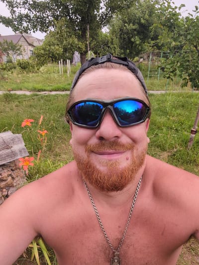 A close-up, selfie-style photograph captures a Caucasian male with a reddish beard and hair pulled back by a black headband. He is wearing dark-lensed sunglasses that reflect a bright blue sky with some tree branches. The man is shirtless, and a silver chain with a pendant is visible around his neck. He is outdoors, with a background of green grass, trees, and some buildings that appear to be in a rural setting, consistent with the village of Perliavka, Ukraine. Several orange lilies are in bloom to the left of the man, and a piece of grey, textured material, possibly stone or concrete, is also present in the foreground. The lighting suggests it is daytime, possibly overcast.