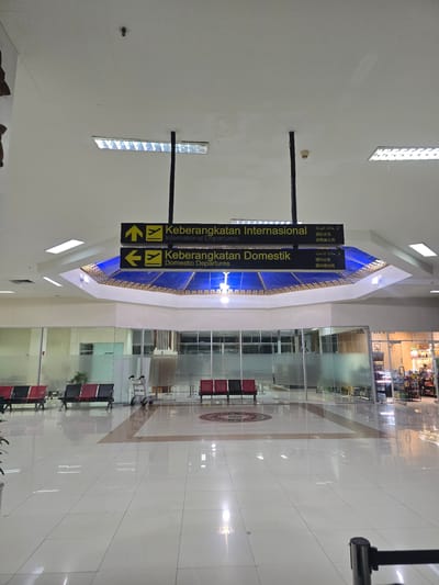 The image depicts the interior of an airport terminal. Overhead, a sign with yellow arrows and text in Indonesian and English directs passengers to "Keberangkatan Internasional" (International Departures) and "Keberangkatan Domestik" (Domestic Departures). Below the sign, a blue ceiling with decorative gold trim is visible. The terminal floor is tiled and highly reflective, showing the overhead lighting.
In the foreground, rows of red and black seats are arranged along a wall of frosted glass. A luggage cart is positioned near the seats. Further into the terminal, more seating and glass-enclosed areas are visible, including what appears to be a shop with shelves stocked with merchandise. The environment appears to be clean and well-lit. The overall scene suggests a typical airport waiting area. The location is specified as Kampungblang, Indonesia.