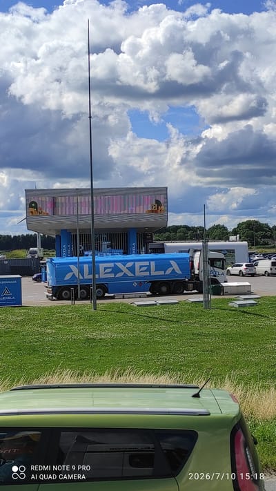 The image shows a blue tanker truck with "ALEXELA" written in large white letters. The truck is parked at a gas station with a modern, cantilevered structure. The gas station appears to be located in Imavere, Estonia, as indicated by the presence of Alexela branding, a well-known Estonian energy company. In the foreground, a green car is partially visible, and behind it, a grassy field stretches out. The sky is filled with dramatic clouds. Several other vehicles, including cars and vans, are parked in the background. The overall scene suggests a typical day at a service station, with the tanker truck likely refueling.