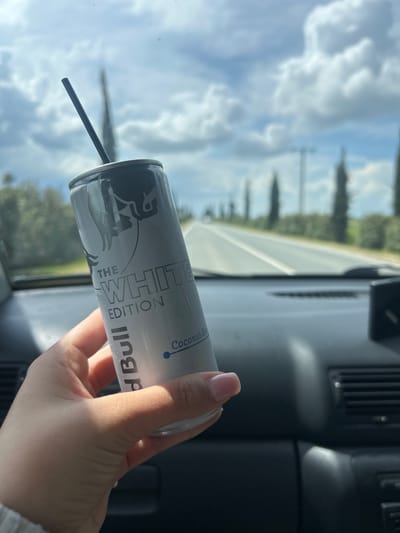 A human hand, displaying light pink nail polish on the visible fingernails, holds a silver and white Red Bull "THE WHITE EDITION" can, which specifies "Coconut-Berry" flavor. A black drinking straw is inserted into the can's opening. The scene is observed from inside a vehicle, with a dark dashboard occupying the lower foreground. Beyond the windshield, a straight asphalt road with white lane markings extends into the distance, flanked on both sides by rows of tall, slender, dark green trees and green foliage. The sky above is bright blue with scattered white cumulus clouds. This environment depicts a road within the vicinity of Xylotymvou, Cyprus.