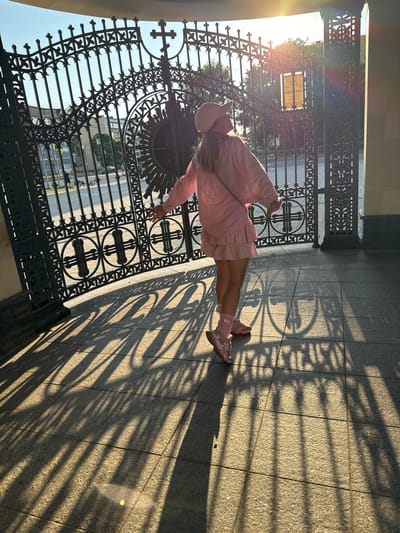A young woman stands in front of a large, ornate black metal gate, her body turned towards the left side of the frame. She is wearing a pink outfit that includes a long-sleeved top, a pleated skirt, a hat, and pink sneakers with light pink accents. Her socks are also pink. The sun is setting, casting long shadows from the gate onto the granite-tiled ground. The light flares strongly from the left, creating lens flare effects. Through the gate, trees and buildings are visible, as well as a street with cars and a motorcycle. A sign with text is attached to the gate. The overall scene suggests a warm, late afternoon or early evening in a public space, possibly a park or a historical site. The mood appears peaceful and serene, with the golden sunlight creating a soft ambiance. The visible text on the sign is in Cyrillic.