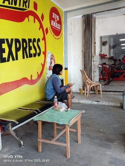 The scene is located in Gegesik, Indonesia at the coordinates -6.536 latitude and 108.432 longitude.  A man wearing a blue denim jacket and dark blue pants sits on a black bench in front of a yellow wall with the words "EXPRESS" in black. The wall is plastered with a red and yellow image that reads "Ayo ke Ahass, Hebat, Mudah & Bergaransi". The man is looking at a phone in his right hand while he holds a bottle of water in his left hand. A small table with a green top and four wooden legs sits in front of the man. A chair is positioned to the right, partially visible. There is an open doorway on the right side of the frame with a small space inside. The interior of the space is cluttered with various objects, including what appears to be a motorcycle tire. The space is bright with light coming in from the front of the building. The floor is grey tile. The space has a small table and a chair. The scene is shot with a slightly tilted perspective. There is no evidence of any illegal activity occurring in the scene.