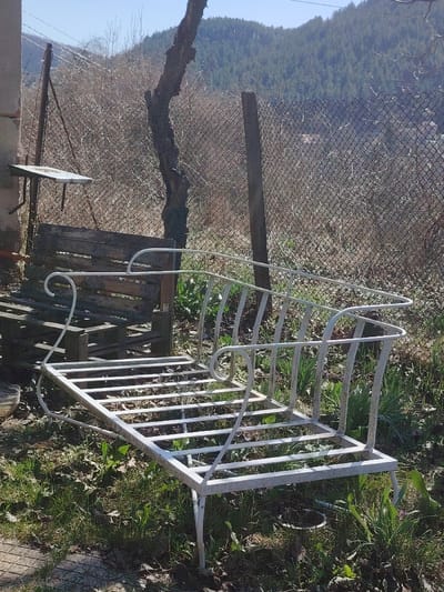 The scene, set outdoors in Vlado Trichkov, Bulgaria, under a clear blue sky, depicts a static, neglected area bordering a forested hillside. In the immediate foreground, a white-painted metal frame, consistent with a garden bench or lounger, lies on patchy grass and exposed dirt, exhibiting surface rust and wear. A small section of light-colored paving is visible in the lower left. To the left, a weathered light-colored wall features a small, white, rectangular shelf affixed to it, with two stacked wooden pallets leaning nearby. A rusty chain-link fence, supported by dark wooden posts and a bare, gnarled tree trunk, spans the mid-ground, separating the foreground from dense, dry, brown underbrush. Beyond the fence, a valley descends, followed by undulating hills densely covered with green coniferous trees. No individuals or animate subjects are present.