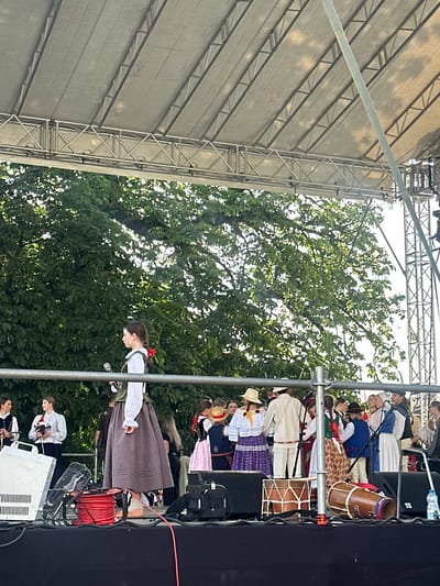 The image depicts an outdoor performance on a stage, likely in the city of Nowy Sącz, Poland. The stage is covered by a large, tent-like structure supported by metal scaffolding. Behind the stage, a dense canopy of green trees is visible against a bright sky.
On the stage, a group of people are gathered, many dressed in traditional folk costumes. In the foreground, a woman in a dark skirt and white blouse with a red ribbon in her hair holds a microphone. To her left, two individuals are visible, one playing a violin. In the background, other performers are dressed in various colorful traditional outfits.
Several musical instruments are placed on the stage, including two drums, one larger and one smaller, positioned in the lower right and lower middle sections of the image respectively. Electrical equipment, including a white speaker or amplifier and a black case, are also present on the stage floor, along with coiled red cables and a black fan. A water bottle sits on a speaker in the far right. The overall scene suggests a cultural event or folk music performance.
