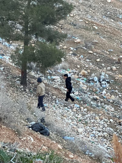 A hillside scattered with trash and small rocks is seen from above. A tall, full pine tree stands on the left. Two figures lie on the slope. One, wearing a black suit, is further up the hill. Another man in a tan jacket and dark pants is below. A black bag or bundled item rests near him. A discarded plastic water bottle sits near a clump of dry bushes.