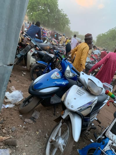 The image shows a crowded street scene in Sokoto, Nigeria, with many motorcycles parked along the side of the road. In the foreground, there are two motorcycles, a blue one and a white one, with a few more out of focus behind them. One man in a yellow shirt stands by the motorcycles. He is wearing a traditional cap. There are many people in the background, some of them walking, some standing, and some sitting on their motorcycles. There are trees lining the road, with dust blowing in the air. The ground is dirt, and there is a lot of litter scattered around. The scene is likely in the Sabon Gari area of Sokoto, where many small businesses and residences are located. The location is in the center of the city, a heavily populated area. The image captures the daily life in a bustling urban center.