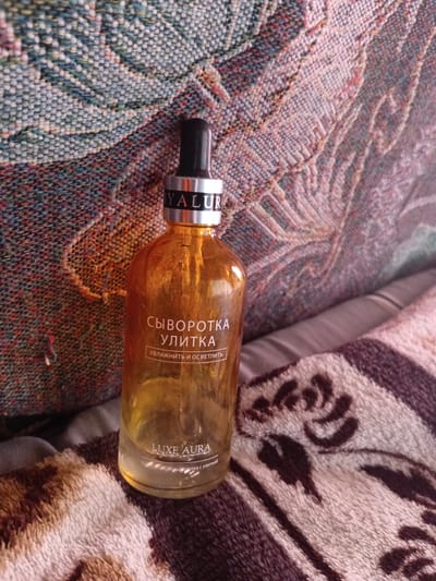A dropper bottle of serum, labeled in Russian as "СЫВОРОТКА УЛИТКА" (Snail Serum) and "LUXE AURA", rests on a textured surface that appears to be a blanket or upholstery with a patterned design. The bottle is made of amber-colored glass and contains a yellow liquid. The dropper cap is black. The background is out of focus, but it suggests a domestic setting. The image does not contain any information that can determine its location in the city of Monastirische, Russia, as it does not depict any identifiable landmarks or features of that city.