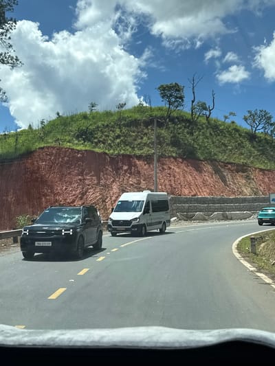 A black SUV with license plate 7BA-217.77 is traveling on a winding road in Thôn 6, Vietnam. A white van is in the lane to the right of the SUV, and a teal car is visible further down the road. The road is bordered by a steep, red-soiled embankment covered in green grass and trees. The sky is blue with scattered white clouds. The scene appears to be a sunny day, likely in the afternoon. The perspective suggests the image was taken from inside another vehicle.