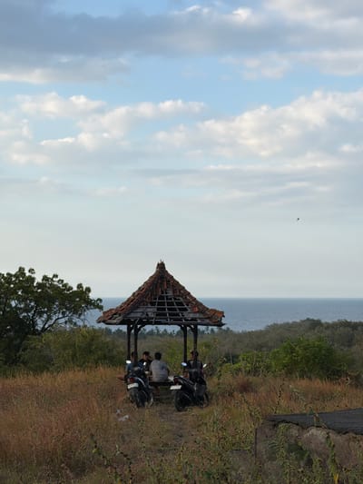 This is a vertical, eye-level, medium shot of a gazebo with a dilapidated roof in Kubutambahan, Indonesia. The gazebo is on a grassy hill overlooking the ocean. Several people are seated inside the gazebo, and motorcycles are parked outside. The sky is partly cloudy, and the ocean is visible in the background. The foreground is filled with dry grass and some green bushes. The overall atmosphere is serene and slightly melancholic due to the weathered appearance of the gazebo.
