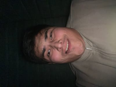 An image taken in Chinabad, Uzbekistan, depicts a male individual lying down, looking directly towards the camera. The subject appears to be of East Asian or Central Asian descent, with dark hair, dark eyes, and a light-to-medium complexion. He exhibits a broad smile, revealing his upper and lower teeth, with slight crinkling around his eyes. He is wearing a light beige or light grey zippered jacket, with a visible metallic zipper pull located on the right side of his chest area. The background behind the individual is a dark, solid color, appearing to be deep green or black fabric, likely upholstery. The lighting is moderate, highlighting the individual's face against the darker background.