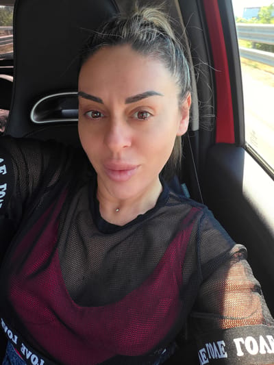 A woman with blonde hair pulled back in a ponytail is taking a selfie in a car. She is wearing a sheer black mesh top over a burgundy bra. The car appears to be a sporty model with a black bucket seat. The background shows a blur of trees and a road, suggesting the car is in motion. The lighting indicates it is daytime. The image does not provide any specific information about the city of Reggio nell'Emilia, Italy.