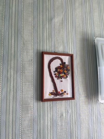 The image shows a framed artwork on a wall with vertical striped wallpaper. The artwork is a braided brown element resembling a pigtail, with a cluster of colorful buttons at the end. There are also a few loose buttons scattered around the artwork and on the wall. The wall features a pale blue and white striped pattern. To the right of the artwork, a sliver of a white object with a metallic frame is visible, possibly another decorative item or part of the room's architecture. The scene is indoors, suggesting a domestic setting. The city of Kovriga, Russia, is not depicted in the image.