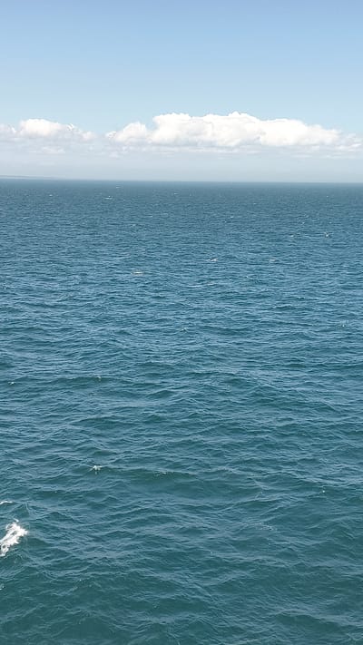 The image depicts a vast expanse of choppy, deep blue ocean under a hazy, pale blue sky with scattered white clouds along the horizon. The sea occupies the majority of the frame, with subtle wave patterns indicating a moderate level of movement. In the distance, a faint, hazy line suggests land or a distant coastline. The sky is clear but muted, with a band of lighter clouds stretching across the upper portion of the image. There are no people, animals, boats, or any other objects visible in the scene, suggesting an open water environment. The lighting is diffuse, typical of an overcast or slightly hazy day. The location is identified as the sea off the coast of the United Kingdom.