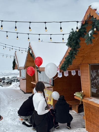 The scene depicts an outdoor winter market or event in Yeghipatrush, Armenia, under an overcast sky. The ground is covered in snow. Several rustic wooden stalls with pitched, snow-laden roofs are arranged in the midground, featuring dark evergreen garlands as decoration. String lights with illuminated bulbs are strung overhead across the scene.
In the foreground, a woman with dark hair styled in two braids, wearing a white long-sleeved top and black trousers, stands with her back to the viewer. She holds multiple red and white balloons, some attached to her person or a phone in her hand. To her left, a person is partially visible, squatting close to the snow-covered ground. To her right, another individual wearing a black puffer jacket and dark pants is also squatting near one of the wooden stalls.
One stall directly in front of the woman displays white and blue bunting with text that includes "YEGHIPATRUSH" and a stylized logo. A menu poster depicting a hot dog is visible on this stall's counter. Another stall to the left has an open service window. A light green, possibly padded, object rests on a wooden bench or shelf attached to the stall on the right. In the distant background, a snowy landscape with hills and a parking area with multiple parked vehicles is visible.