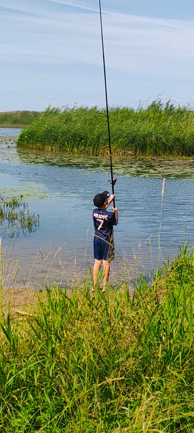 A young person wearing a dark blue shirt with "MBAPPE" and the number "7" on the back, and dark shorts, stands at the edge of a body of water. They are holding a fishing rod, with the tip extended upwards. The water is calm with some lily pads visible. Tall green reeds grow along the bank and in the distance. The sky is blue with some wispy clouds. The scene appears to be a natural outdoor setting, likely a river or lake, and is set in the city of Malaya Listovka, Russia.