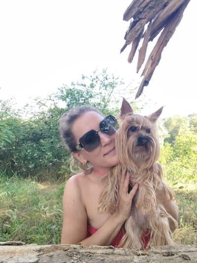 A woman with blonde hair pulled back and wearing large, dark sunglasses is holding a small, tan Yorkshire terrier dog. The dog is being held close to her face, and she is smiling slightly. The woman is wearing a pink top, and a pink necklace is visible. They are outdoors, with a background of lush green trees and foliage. A piece of weathered wood is in the foreground, and a larger, broken piece of wood is seen in the upper right corner of the image, with a bright sky peeking through. The lighting suggests it is daytime.