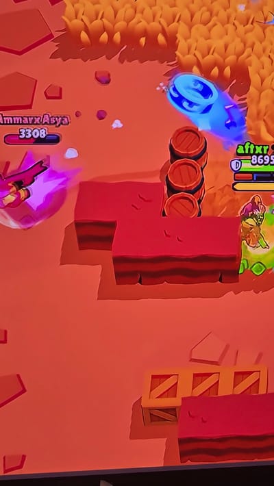 This image depicts a screenshot from a video game, not a real-world location in Genk, Belgium. The scene presents a top-down, isometric view of a digital arena map with a reddish-orange ground texture.
In the upper left quadrant, a character identified as "AmmarXAsya" with 3308 health is initiating an action, characterized by a bright pink energy emission directed downwards and to the right. In the upper right quadrant, a blue, ringed, barrel-shaped entity, identified as "aftxr" with 8695 health and an active 'D' icon, is shown in rapid motion, leaving a blue trail. Below "aftxr," a third character, partially visible with green and brown elements, stands near a red block obstacle.
Environmental elements include patches of tall yellow-orange foliage along the top edge of the frame. Multiple dark red, L-shaped block obstacles are arranged centrally. Three brown wooden barrels with black bands are stacked vertically behind a red block obstacle. In the lower right corner, a yellow wooden crate with orange diagonal strapping is visible, next to a partially obscured similar crate. Small red and white fragments are scattered across the ground. All visible structures cast distinct shadows on the reddish-orange terrain.