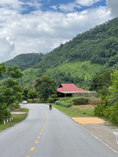 A paved road curves through a lush green mountainous landscape in Thuận Hòa Commune, Vietnam. On the right side of the road, a large red-roofed structure stands, partially obscured by trees. In front of the structure, a field of dried corn is spread out on the ground. Further down the road, a person rides a motorcycle. To the left of the road, a white car is parked, and a few small white posts with red tops line the roadside. The sky is cloudy with patches of blue visible. The mountains are densely covered in trees.