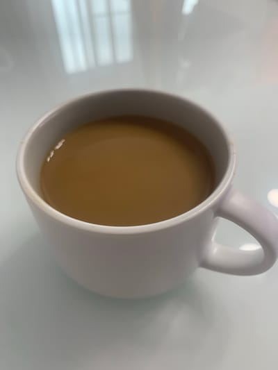 The media file is a close-up, overhead shot of a white mug filled with a light brown liquid, likely coffee or tea with milk, resting on a white, reflective surface. The mug is tilted, with the handle visible on the right side of the frame. The liquid inside the mug has a smooth surface, with a few subtle highlights reflecting light. The background is predominantly a soft white, with subtle shadows and reflections suggesting an indoor environment, possibly a table or countertop. The lighting appears to be artificial, with soft illumination creating gentle gradients and reflections on the surface and the mug. There is no visible text in the image. The scene depicts a simple, everyday moment, possibly a pause for a beverage. The overall mood is calm and neutral. The location context provided is Bugry, Russia, but there are no specific visual cues in the image to confirm this location.
