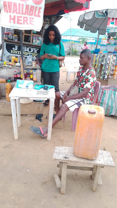 The image shows a roadside business scene in what appears to be Warri, Nigeria. A woman with dark hair and wearing a green shirt is looking at her phone, standing near a stall. A man wearing a patterned shirt and shorts is seated, looking off to the side. A small white table displays a POS machine and other items. Behind them is a makeshift stall displaying various goods, including drinks and engine oil. A sign reading "AVAILABLE HERE" and "J BOY ENTERPRISES" is visible. An orange jerrycan sits on a wooden stool next to the seated man. The ground is dirt and gravel.