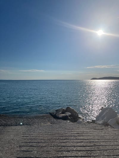 The image depicts a serene seascape at Ποτός, Greece, on a clear, sunny day. In the foreground, a concrete ramp with horizontal indentations leads down to the water's edge, where large rocks and boulders are situated. The sea, a deep blue, stretches towards the horizon, reflecting the bright sunlight in a dazzling display of sparkles. In the distance, a hilly coastline is visible under the expansive sky, which is partially obscured by the sun's glare and a few wispy clouds. There are no people or animals visible in the frame. The overall impression is one of tranquility and natural beauty, characteristic of a coastal Mediterranean setting.