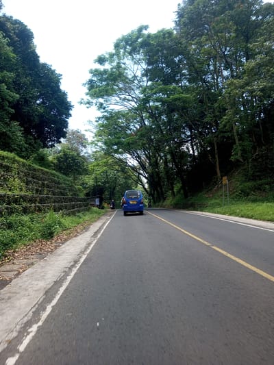 A road curves through a heavily wooded area. A blue minivan travels away from the camera on the right side of the road, straddling the yellow center line. A motorcyclist trails behind the van. A wall composed of tiered stone lines the road on the left, with trees on both sides of the road creating a canopy overhead. Based on metadata, the location is Nawng Hkar, Myanmar.