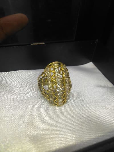 A gold-toned ring, featuring an ornate, elongated, open-work head set with multiple rows of small, round, clear stones, is centered on a white, satin-like fabric surface. This fabric functions as the lining of a dark-colored jewelry box or display case, indicated by dark borders framing the material. The upper background consists of a dark, reflective surface, likely the interior of the box lid, which shows a partial, blurry reflection of a light brown human digit in the upper-left quadrant. The scene is illuminated from the front, highlighting the ring's details and the fabric's texture. The image provides no visual information to identify a specific location within the city of Liège, Belgium.