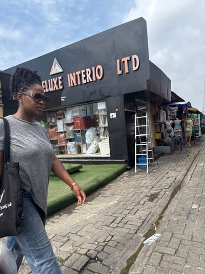 The image depicts a daytime street scene at 796 Ajah-Badore Road, Ajah, Nigeria. In the foreground, a woman of African descent walks from left to right, wearing large dark sunglasses, a grey short-sleeved top, blue jeans, and a gold-colored wristwatch on her left wrist. She carries a black tote bag on her left shoulder. Her dark hair is styled in braids or twists.
The prominent structure in the midground is a commercial building with a modern, dark grey or black facade, featuring the name "DELUXE INTERIO LTD" in raised, orange-colored letters. The address "796" is visible below the name. Large display windows showcase various home furnishings and textiles. Artificial green turf lines the ground directly in front of the building's windows. To the right of the main entrance, an aluminum stepladder leans against the building, with a blue plastic bucket at its base. An air conditioning unit is mounted on the wall beside the ladder.
To the right of the "DELUXE INTERIO LTD" building, a series of less structured market stalls with tarpaulin awnings are visible, displaying clothing on mannequins. Several individuals are present in this area, interacting with the stalls. The ground consists of grey interlocking concrete pavers, which appear worn and contain a narrow concrete drainage channel with standing water. Scattered debris, including a piece of paper, is visible on the pavement. The sky above is partially cloudy.