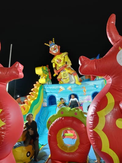 The image shows a brightly colored, inflatable children's bounce house with a large, yellow Monkey King figure sitting atop. The bounce house also features a yellow and green inflatable dragon. Several children are inside the bounce house. Two large, red inflatable dragons flank the bounce house in the foreground. The scene appears to be at night, against a dark sky. The location is Blitaran, Indonesia.