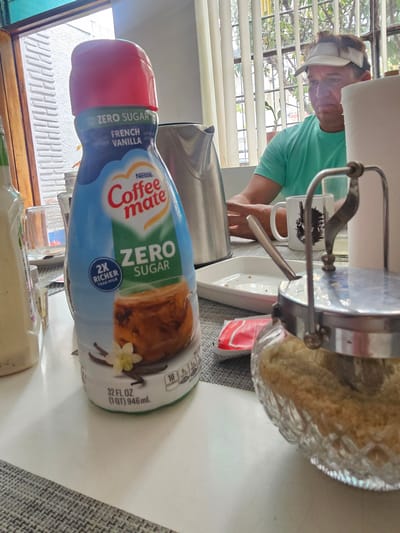 A residential interior in Magdalena del Mar, Peru, features a partial view of a woman with a visor wearing a green shirt, standing near a window with closed blinds. Adjacent to her on a countertop are a mug, a sugar dispenser with brown sugar, and a half-empty bottle of Nestle Coffee-mate Zero Sugar French Vanilla creamer. A silver kettle is visible behind the creamer bottle. The countertop is a light gray or white color, and the surroundings appear to be part of a kitchen or dining area.