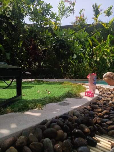 The image depicts an outdoor scene in Hua Hin City Municipality, Thailand, near a pool area. A young girl with blonde hair is visible near the edge of the pool, which is bordered by smooth, dark-colored pebbles. Beside her, on the concrete edge, are a pink plastic hair clip and a pink tumbler decorated with a blue character. Beyond the pool edge, there is artificial turf with scattered leaves and flowers, and a black lounge chair is partially visible on the left side. The background features lush tropical foliage, including plumeria trees with white flowers and various palm trees, obscuring the view of any structures. The scene is bathed in sunlight under a clear, blue sky.