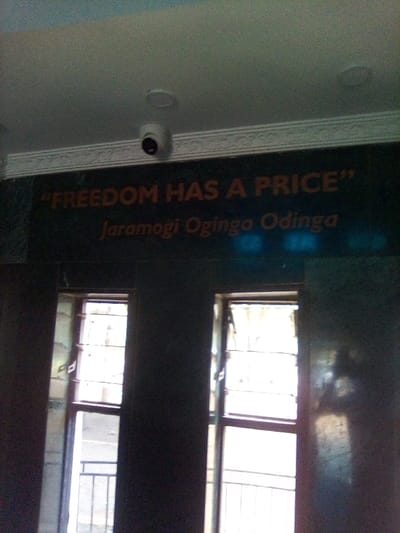 The scene depicts an interior view within a building, specifically in South Sakwa ward, Kenya. A dark wall occupies the majority of the frame, featuring the quote "FREEDOM HAS A PRICE" in orange capital letters, attributed below to "Jaramogi Oginga Odinga" in smaller orange text. Above this wall, a light-colored ceiling with recessed lighting fixtures is visible, from which a white, dome-shaped CCTV camera is mounted, pointed downwards. A decorative white molding separates the ceiling from the dark wall.
Below the quote, two tall, vertical windows are symmetrically positioned within the dark wall. Each window has light-colored frames and is divided into multiple panes, with the top section appearing slightly open or ajar. Metal security bars are visible through the lower sections of both windows, indicating an exterior beyond. Bright natural light floods through the windows, creating glare on the dark wall surfaces adjacent to them and obscuring precise details of the outdoor environment, though faint outlines of exterior structures or ground are discernable. No individuals are present in the image.