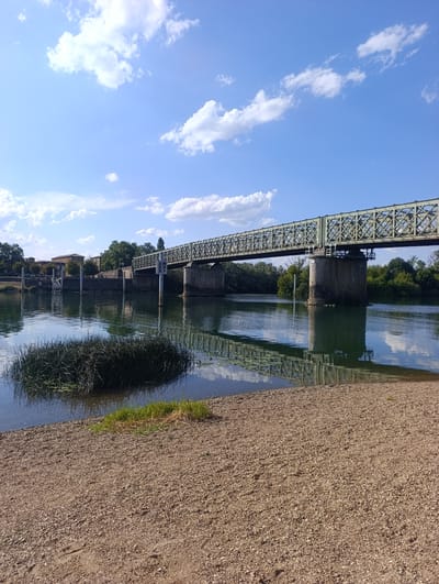 An eye-level, long shot captures a metal truss bridge spanning a calm river under a bright blue sky with scattered white clouds. The bridge, painted a muted green, is supported by stone pillars and shows a decorative lattice design. The river's surface reflects the bridge and the sky, with a small island of reeds in the foreground. The riverbank is covered in small stones and gravel. To the left of the bridge, buildings and trees are visible on the far bank. A navigational marker with directional arrows stands in the river near the bridge. The scene is set in Guéreins, France, near the Saône river.